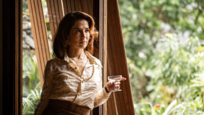 Marjorie Estiano como Ângela Diniz, de camisa de seda e colar dourado, segura taça de bebida em cena de série da HBO Max