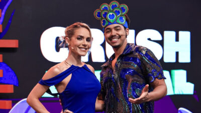Monique Alfradique de vestido azul e Pedro Ottoni com camisa de paetês e adereço de cabeça posam juntos no palco de Crush Animal