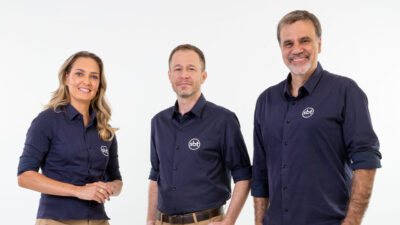 Nadine Basttos, Tiago Leifert e Mauro Beting sorrindo para a câmera, usando camisas sociais azuis escuras com o logo do SBT