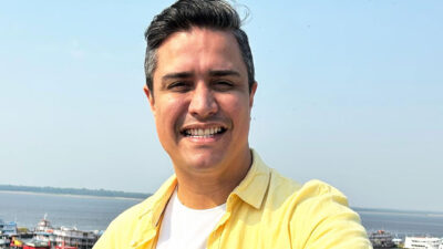 Jornalista Paulo Carneiro sorrindo com camisa amarela e cabelo penteado para o lado em uma paisagem de rio e céu azul