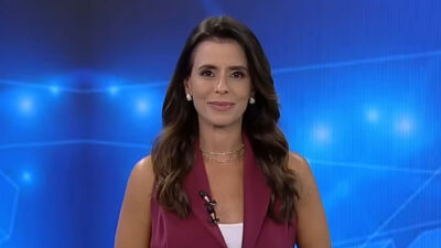 Jornalista Renata Caetano sorrindo, usando blazer regata vinho e colares discretos no estúdio de telejornal da Record News
