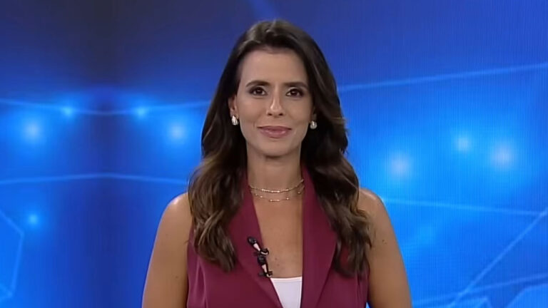 Jornalista Renata Caetano sorrindo, usando blazer regata vinho e colares discretos no estúdio de telejornal da Record News