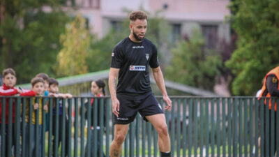 Jogador Renato Soares em campo de futebol usando uniforme preto, com crianças assistindo atrás da grade
