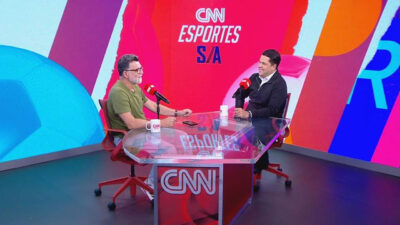 Ricardo Rocha e João Vitor Xavier no estúdio da CNN Esportes S/A sentados em cadeiras vermelhas em frente à bancada transparente