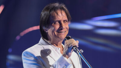 Cantor Roberto Carlos sorrindo de olhos fechados, usando terno branco enquanto canta no microfone em show