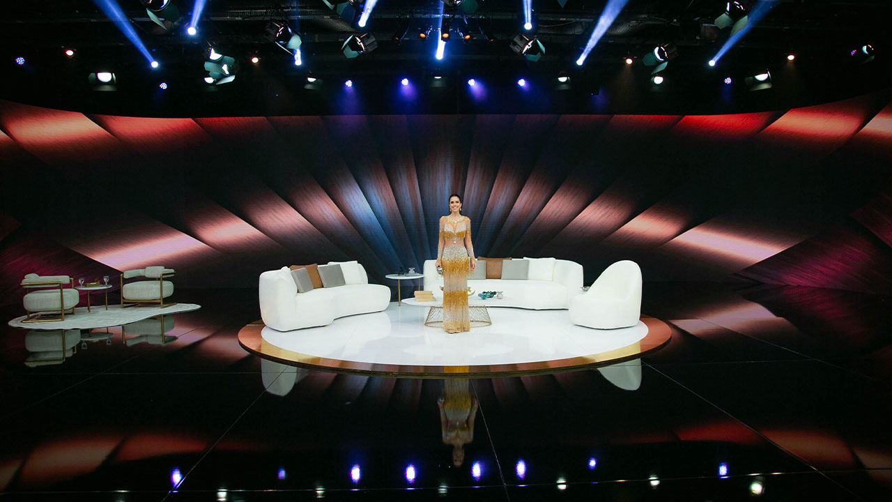 Daniela Albuquerque no novo cenário do Sensacional, vestindo um longo vestido dourado, com sofá branco no centro do palco