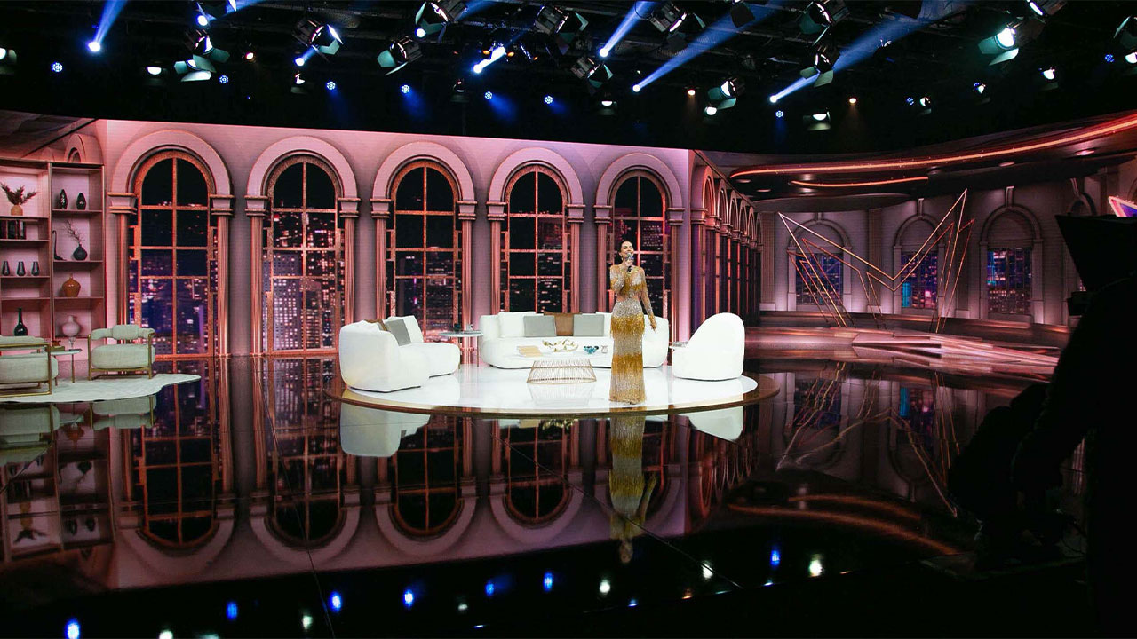 Cenário do programa Sensacional da RedeTV! com lustres e janelas com vista noturna, e um sofá branco no centro do palco