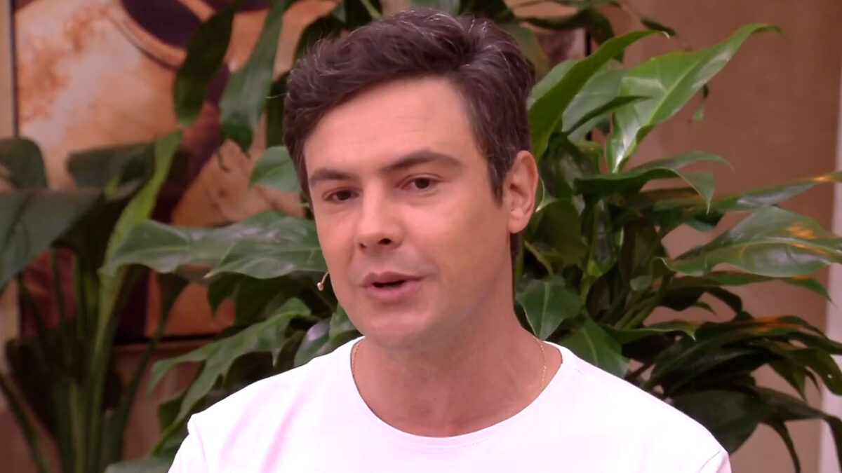 O ator Sérgio Guizé com camiseta branca e corrente dourada fala durante entrevista em um cenário com plantas verdes ao fundo