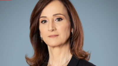 Thais Herédia, com cabelo castanho e blazer preto, com olhar sério em fundo azul