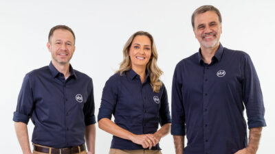 Tiago Leifert, Nadine Basttos e Mauro Beting lado a lado, sorrindo e usando camisas azuis do SBT em estúdio branco