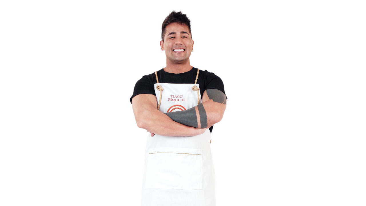 Tiago Piquilo, sorrindo, de braços cruzados, vestindo camiseta preta e avental MasterChef com o braço tatuado à mostra