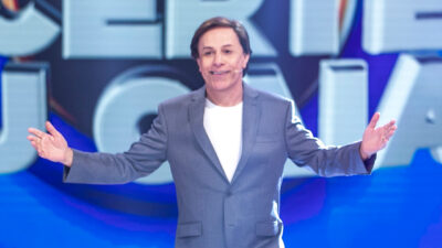 Apresentador Tom Cavalcante no palco do Acerte ou Caia! da Record com terno cinza, gesticulando com os braços abertos