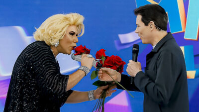 Apresentador Celso Portiolli de terno preto e personagem Webe de peruca loira trocando rosas vermelhas no palco do SBT
