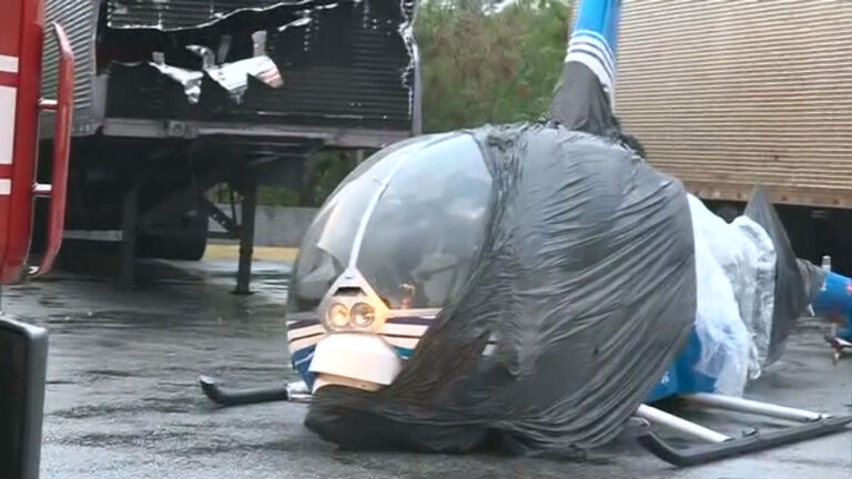 Helicóptero da Band com cabine coberta por plástico preto no asfalto após pouso de emergência e caminhão baú danificado ao fundo da imagem