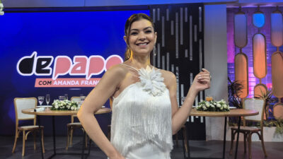 Amanda Françozo sorri com vestido branco de franjas e flor no estúdio do programa De Papo com Amanda Françozo na TV Aparecida
