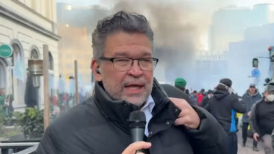 Américo Martins segura microfone da CNN Brasil durante cobertura de protesto com fumaça e manifestantes ao fundo em rua urbana