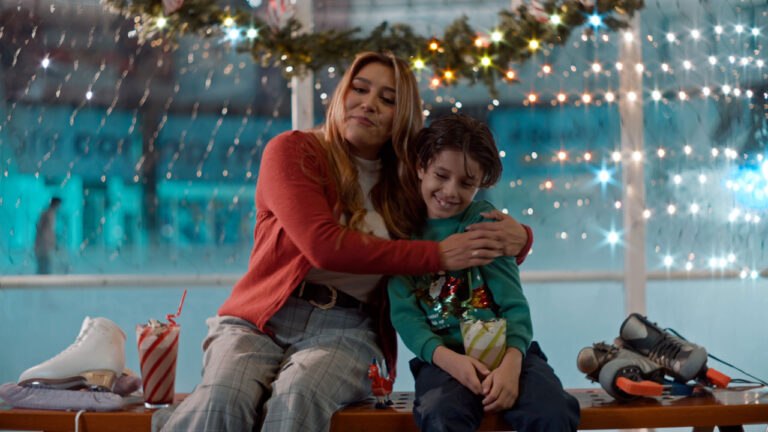 Mulher de vermelho abraça menino de verde sentados em banco com patins e luzes ao fundo em cena de Arquitetando o Amor no Natal