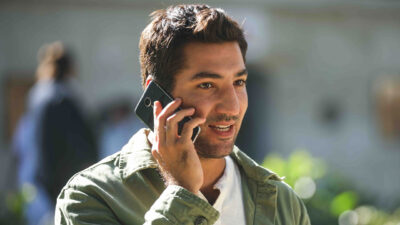 Berkay Ates como Ozan Akinsel sorri enquanto fala ao celular vestindo camisa verde sobre camiseta branca em cena da novela Chamas do Destino