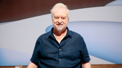 Humorista Beto Silva sorri de camisa azul marinho e barba branca em estúdio de TV com painel abstrato azul e branco ao fundo