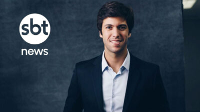 Caio Coppolla de terno escuro e camisa clara posa sorrindo ao lado do logotipo do SBT News em fundo azul