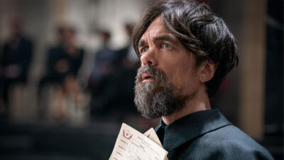 Peter Dinklage com barba e cabelo desgrenhado, em close, olhando para o lado, segurando um papel branco na mão