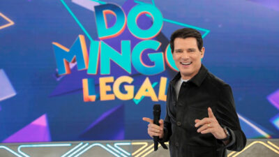 Celso Portiolli sorrindo com jaqueta preta e microfone na mão no palco do Domingo Legal, com o logotipo do programa exibido ao fundo