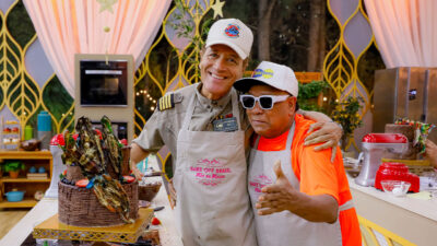 Comandante Hamilton e Liminha sorriem abraçados usando aventais do Bake Off Brasil ao lado de um bolo de chocolate no cenário do SBT