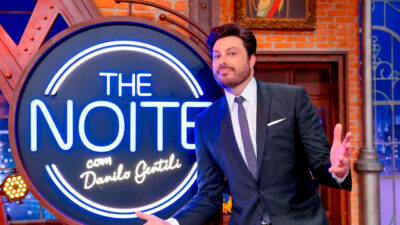 Danilo Gentili de terno abre os braços ao lado do logotipo luminoso do The Noite no cenário do programa de entrevistas do SBT