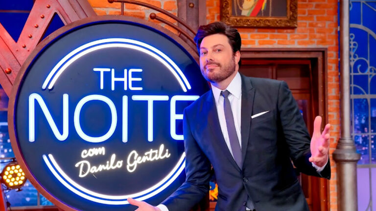 Danilo Gentili de terno abre os braços ao lado do logotipo luminoso do The Noite no cenário do programa de entrevistas do SBT