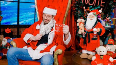 Danilo Gentili sorri vestido de Papai Noel com caneca vermelha sentado em poltrona no cenário decorado do The Noite no SBT