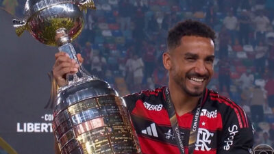 Danilo Luiz sorrindo, vestindo a camisa do Flamengo e segurando o troféu da Copa Libertadores no meio do campo com estádio lotado