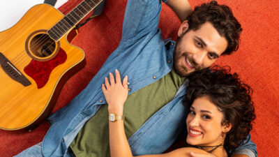Diego (Gustavo Mioto) e Cindy (Maya Aniceto) deitados e sorrindo com violão ao lado e fundo vermelho