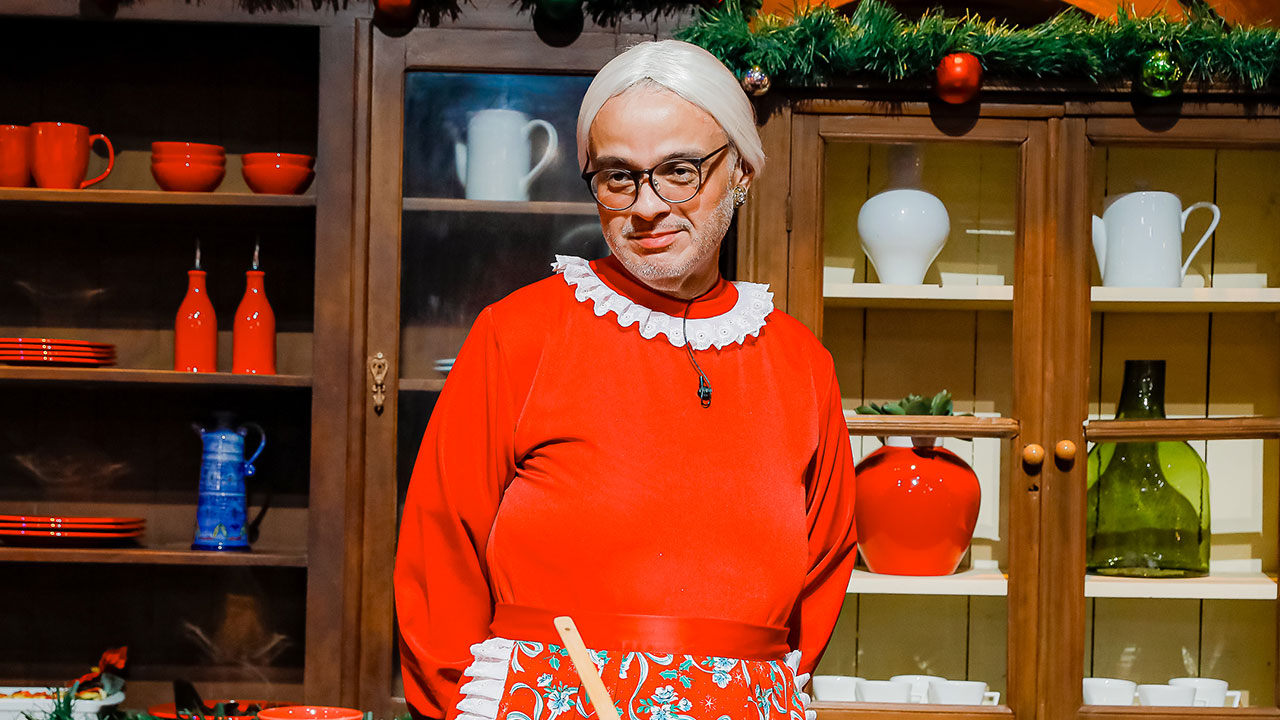 Diguinho Coruja veste roupa de Mamãe Noel com peruca branca e óculos em cenário de cozinha durante especial de Natal do The Noite