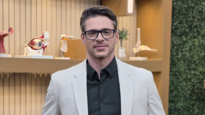 Dr Rodrigo Gurgel de blazer bege e camisa preta no cenário do programa Você Bem Melhor com modelos anatômicos em prateleira ao fundo