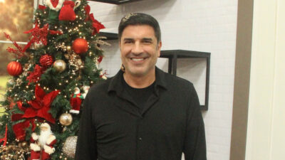 Edu Guedes sorri vestindo camisa preta ao lado de uma árvore de Natal decorada com bolas vermelhas e bonecos de Papai Noel