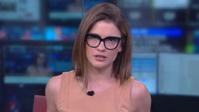 Elisa Veeck de óculos e blusa marrom claro olha séria para a câmera durante a apresentação de telejornal no estúdio com telas ao fundo