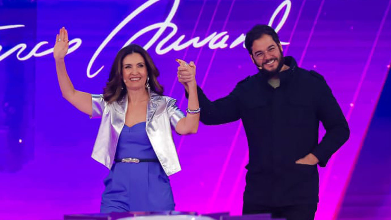Fátima Bernardes sorri de mãos dadas com Túlio Gadelha no palco do Programa Silvio Santos com fundo roxo