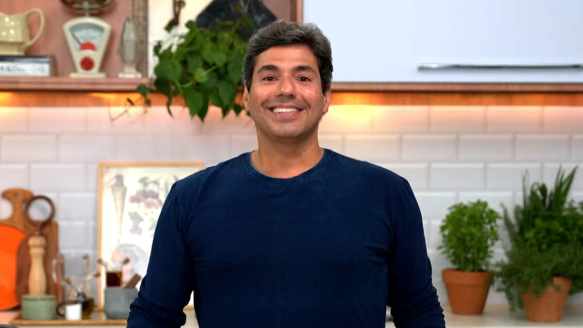 O chef de cozinha Felipe Bronze sorrindo com camisa azul em estúdio de TV com azulejos brancos e plantas na decoração do cenário