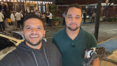 Selfie de Felipe Sousa, sorrindo e usando um moletom cinza, ao lado de Gianvitor Dias, que usa uma camisa polo verde escura, segurando um celular em um estabilizador. Eles estão em uma rua à noite, com um grupo de pessoas e um restaurante ao fundo