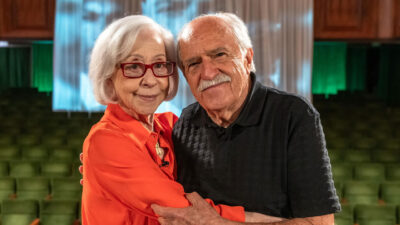 Fernanda Montenegro de blusa laranja abraçada a Ary Fontoura de camisa polo preta no cenário do Conversa com Bial na TV Globo