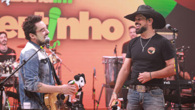 Fernando e Sorocaba sorriem no palco da Band com Fernando tocando violão e Sorocaba de chapéu usando microfone