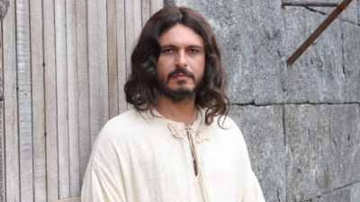 Flávio Rocha como Jesus de cabelos longos e túnica clara olha sério para a frente em cena do filme Milagres de Jesus na Record