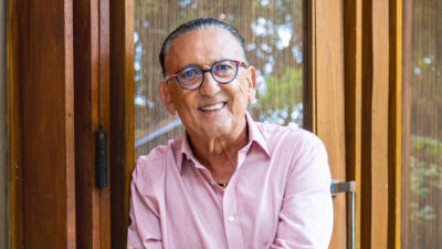 Galvão Bueno sorri usando óculos e camisa rosa clara enquanto posa encostado em uma estrutura de madeira e vidro com reflexo de árvores ao fundo