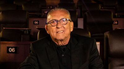 Galvão Bueno usa óculos e terno preto sentado em poltrona de cinema numerada durante o lançamento de seu documentário no Globoplay