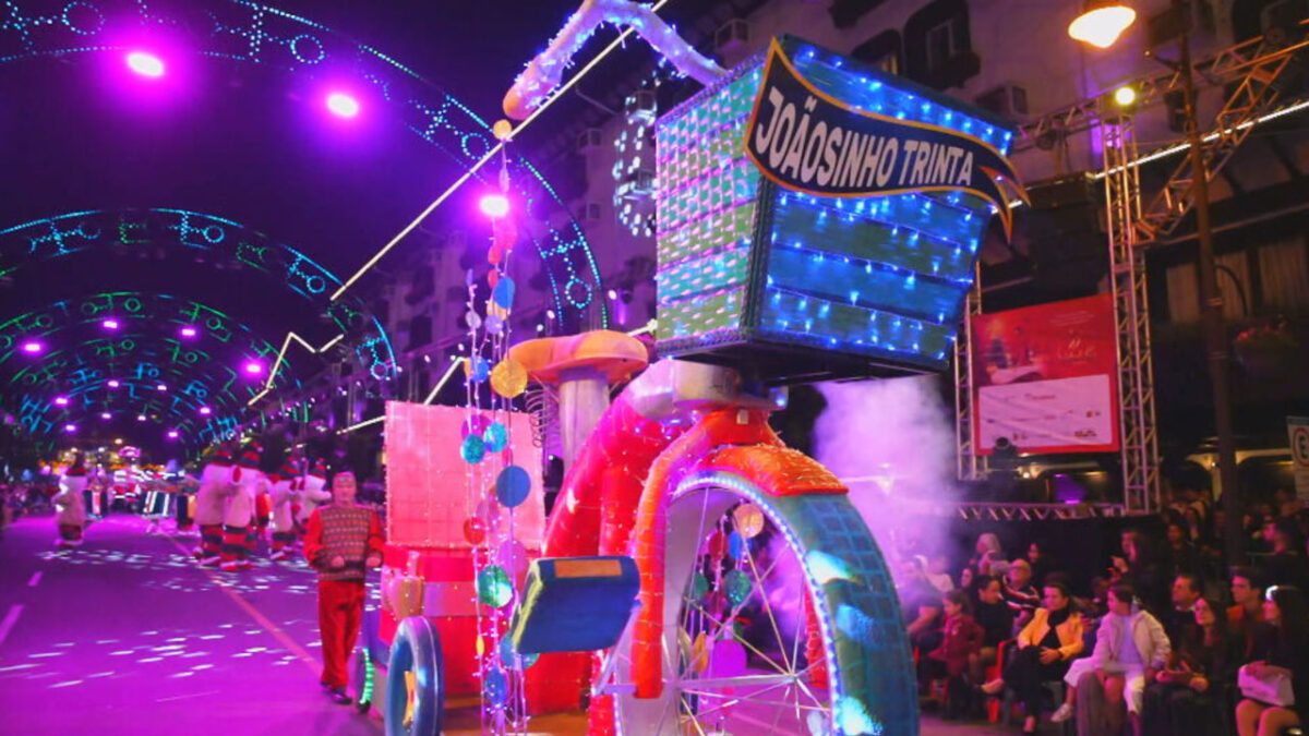 Carro alegórico em formato de triciclo colorido e iluminado com o nome Joãosinho Trinta durante desfile do Natal de Gramado