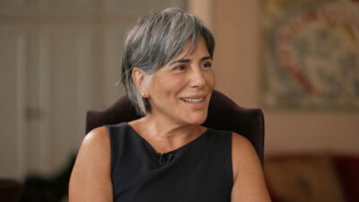 Gloria Pires sorrindo, em um plano médio, usando um vestido preto e cabelo grisalho curto em entrevista