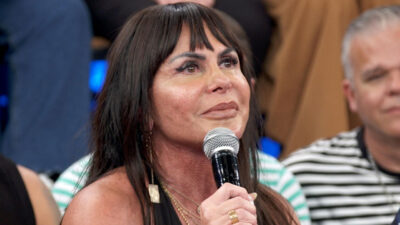 Cantora Gretchen fala ao microfone com cabelos soltos e joias douradas em estúdio de TV com homem de camisa listrada ao fundo