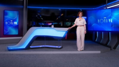 Thais Herédia sorrindo em pé ao lado da bancada futurista iluminada do estúdio do telejornal Hora H