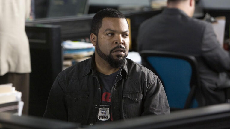 Ice Cube como James Payton no filme Policial em Apuros usa camisa preta e distintivo policial em uma delegacia com mesas ao fundo
