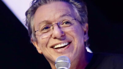 J.B. de Oliveira Boninho sorrindo com óculos de aro fino e microfone próximo ao rosto em evento